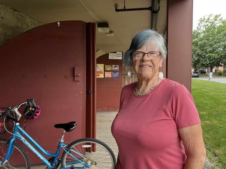 A woman with short, grey hair stands smiling in a pink t-shirt. Behind her is a blue bicycle and a row of rust-coloured casemate doors.  