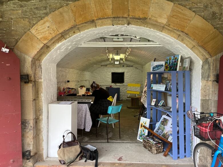 A person in a black shirt sits down at a table inside a brick casemate. Around them, are several paintings and arts and crafts.