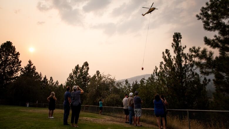 People take pictures and look up at a yellow helicopter in the sky, which is covered by haze and wildfire smoke.