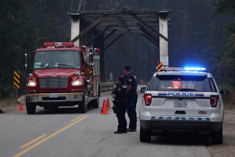 Uniformed police stand next to a police vehicle on a roadway in front of a bridge and a fire truck.