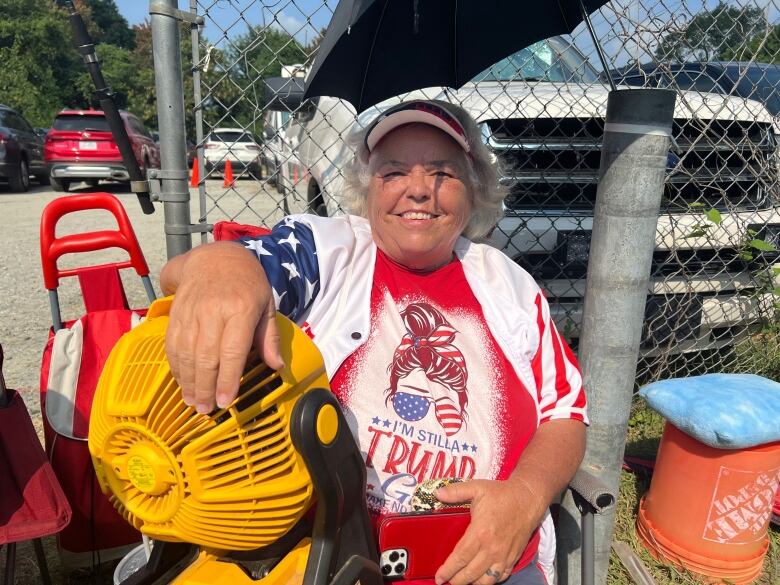 A smiling woman wearing a pro-Trump t-shirt sits with her hand on a yellow portable fan under an umbrella. 