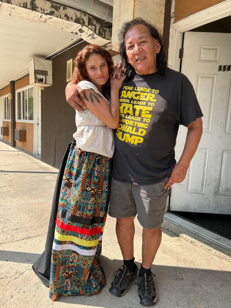 A couple stand together in the sun outside a rundown motel.