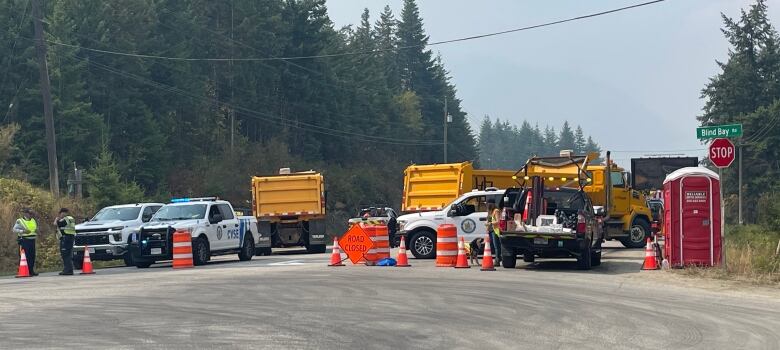 Two people in high visibility vests stand beside several vehicles blocking a highway.