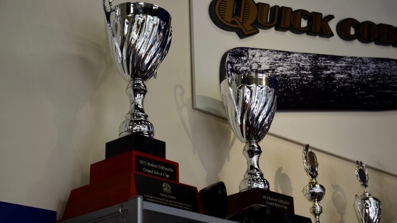 Two large silver trophies shaped like goblets with swirls on the cup sit in a row on a high shelf next to smaller silver trophies.