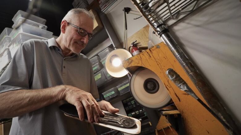 A man with short grey hair and a grey golf shirt holds a piece of shoe sole near a sanding wheel.