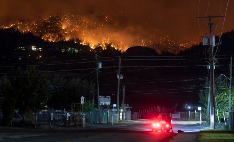 Two motorcyclists stop at an intersection and watch a wildfire burning on a nearby mountain ridge.