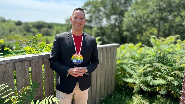 A man wearing a black blazer over a red shirt and a rainbow necklace stands smiling.