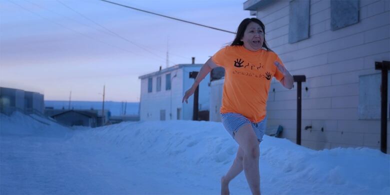 Still frame from the film Tautuktavuk (What We See). Lucy Tulugarjuk runs barefoot through the snow, wearing an orange t-shirt and a distressed look on her face.