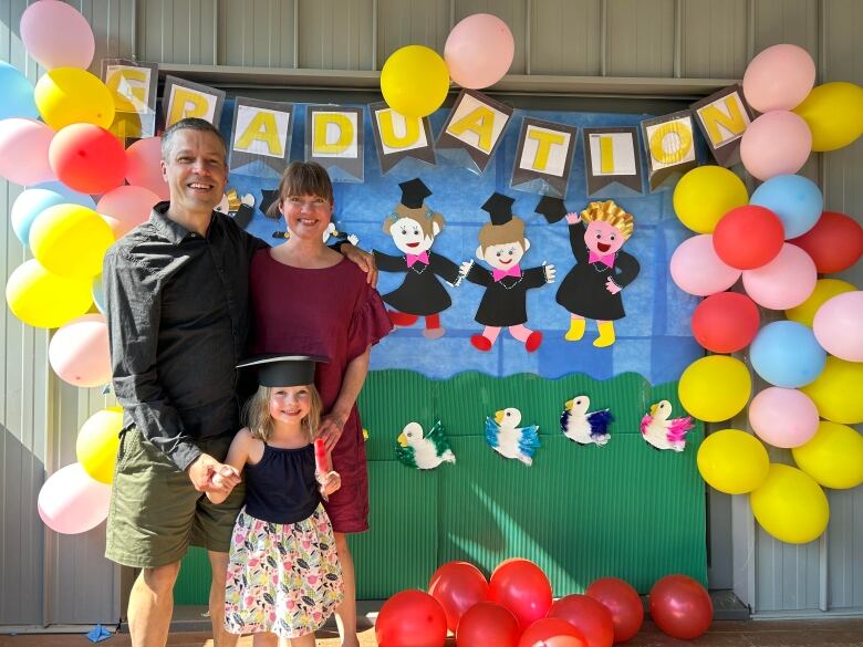 A smiling couple with a young children stand in front of a colourful mural that says 'graduation.'