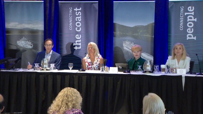 Four people sat at a long banquet table covered with a white cloth and a black curtain that hangs down and hides the lower half. Each person has their own microphone. Behind them are blue folded drapes that are interspersed with signs that say Connecting the Coast.