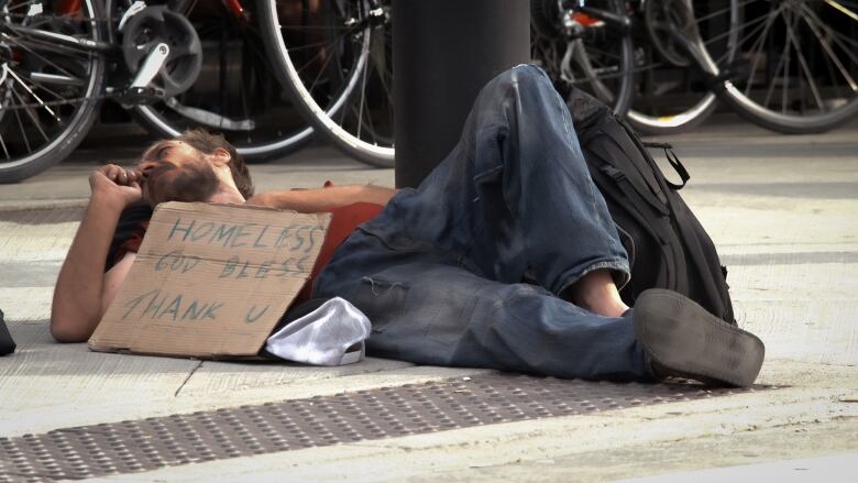 A man suffering from homelessness is lying down on the sidewalk. His sign reads, 