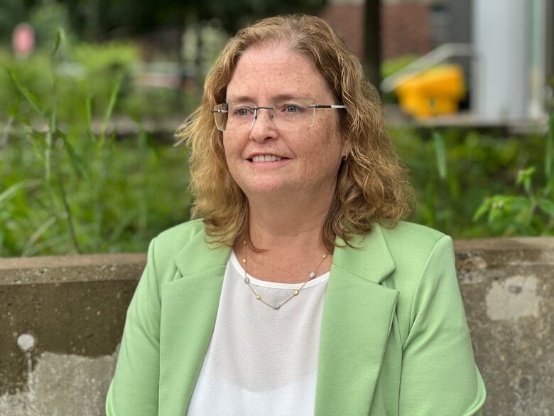 A woman with a light green jacket, blonde curly hair and glasses stands in front of a cement wall with grass behind it.