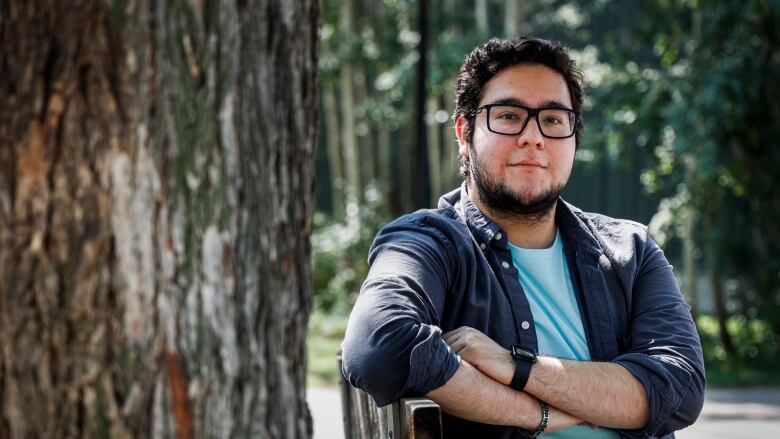 A young man is sitting and looking at the camera. Behind him there are trees. He's wearing a light blue T-shirt and a navy button down over top. 