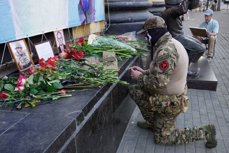 A man wearing a face mask and military uniform kneels in front of a memorial with flowers and the portraits of two men.