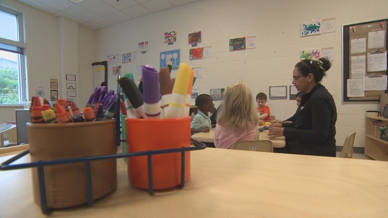 And early childhood educator and her students sit around a table in a daycare centre. There are crayons and markers in the foreground.