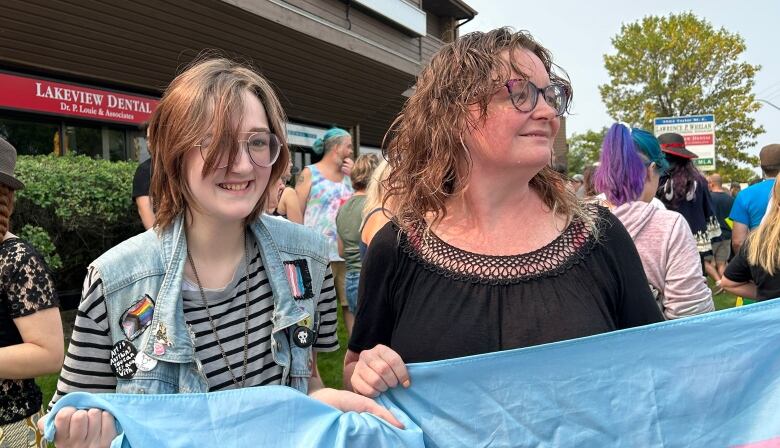 A boy and his mother are holding a transgender pride flag. A crowd of people are behind them, in front of an office building.