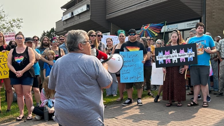 A woman with short grey hair, wearing a grey t-shirt, is speaking into a megaphone to a crowd of people holding signs and flags.