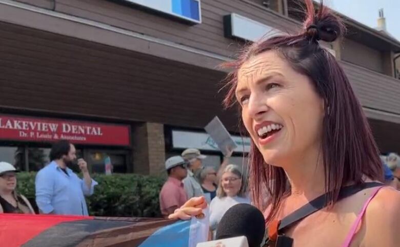 A woman with purple hair and yellow top is holding a pride flag and speaking to someone holding a microphone with the CBC logo on it. A crowd of people are behind her.