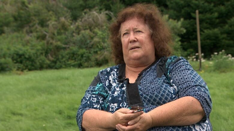 A woman wears a blue-patterned top. She has auburn hair and sits in her yard. 