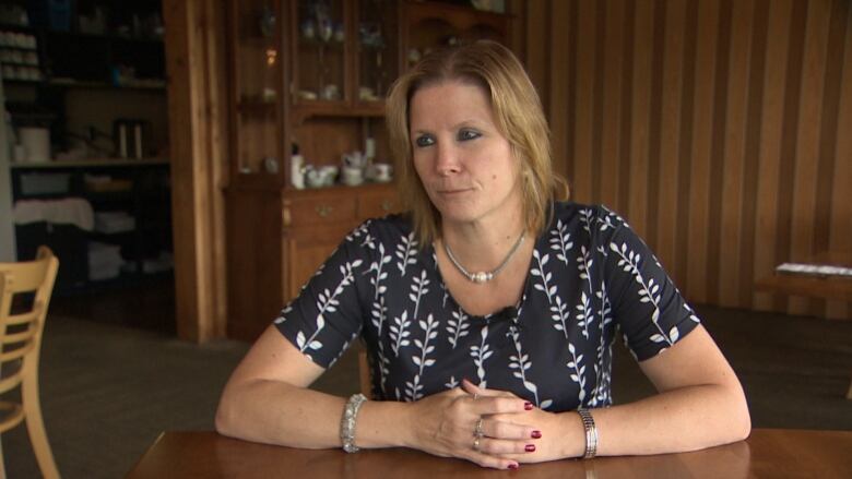 A women wears a black and white shirt. She sits with her hands together at a brown wooden table. 