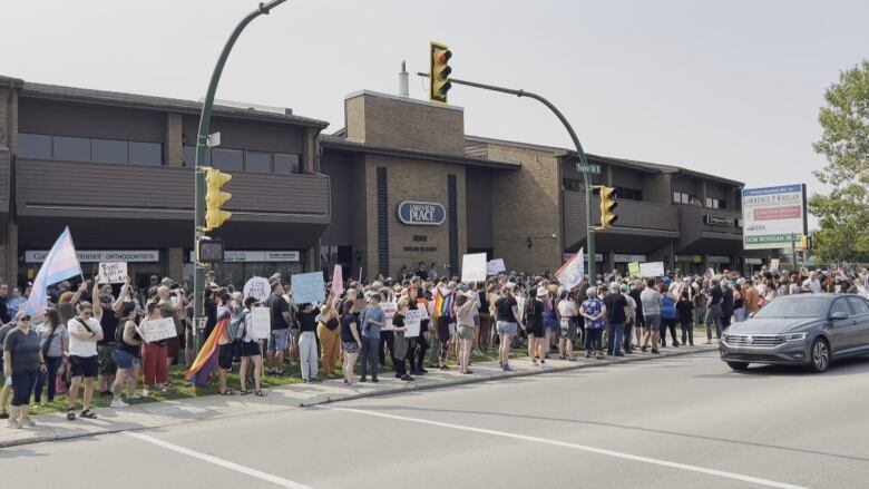 A crowd of people, some of whom are holding pride flags and handwritten signs, are standing on the lawn in front of a brown, brick office building. The building is located at a three-way intersection; the street lights on the same lawn as the crowd. A grey Volkswagen car is driving through the intersection from the right of the photo. 