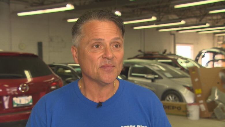 A man is standing inside an autobody garage, with vehicles in the background.