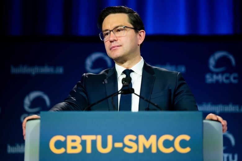 A man in glasses, suit and tie speaks at a podium.