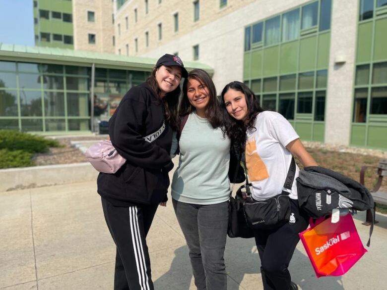 Three young women with brown hair and light skin pose smiling, arm-in-arm, in front of a dorm building.