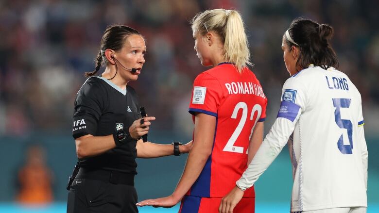 A soccer referee wearing a black uniform is talking to two players one in red and one in white at the 2023 Women's World Cup. 