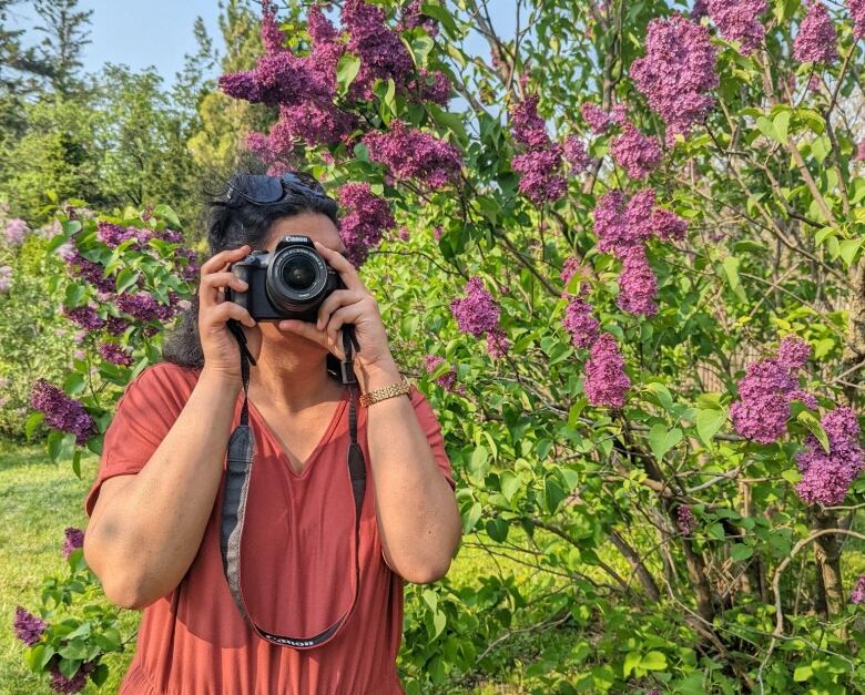 A woman looks through a camera in front of colourful flowers.