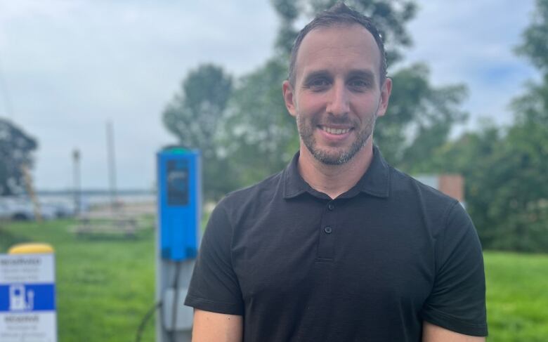 A man stands in a black polo shirt against a backdrop of electric vehicle chargers.