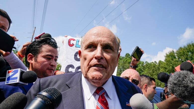 A man in a suit and tie speaks into microphones and recording devices held by reporters in an outdoor photo.