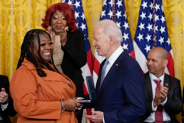 A woman is presented with a item by a man in a suit and tie in a room with an American flag in the background.