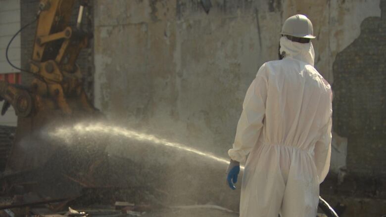 A person wearing a hazardous materials suit is spraying water from a hose. An excavator is working in the background.