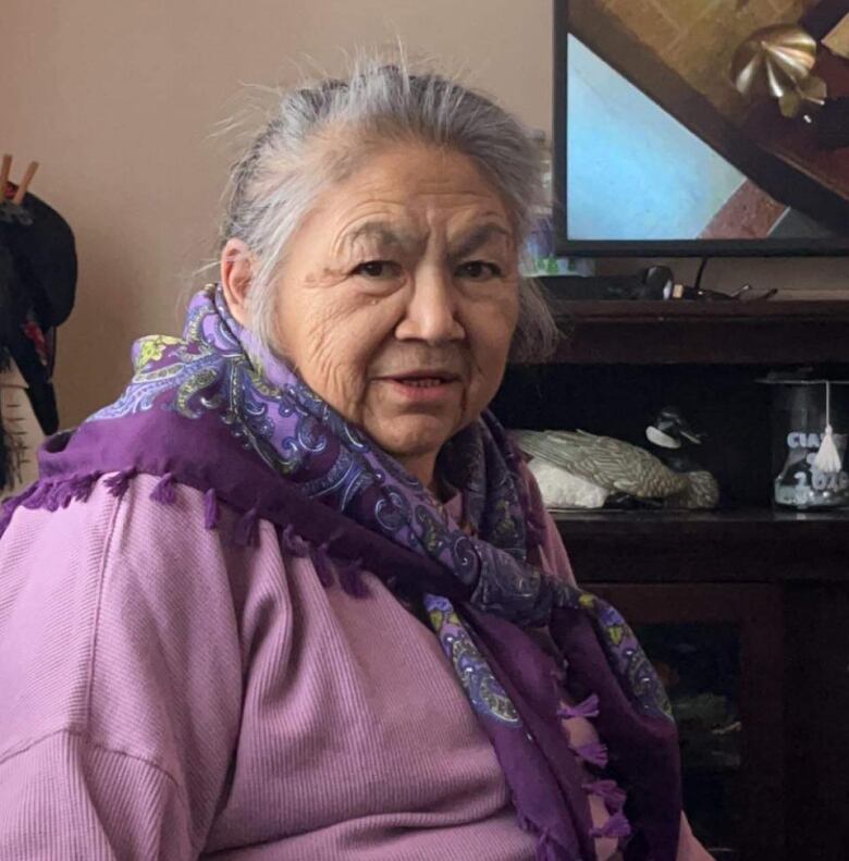 An elder dressed in purple with a googkum scarf looks into the camera.