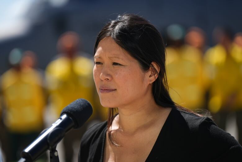 A woman is pictured at an outdoor news conference, with firefighters wearing yellow visible in the background.