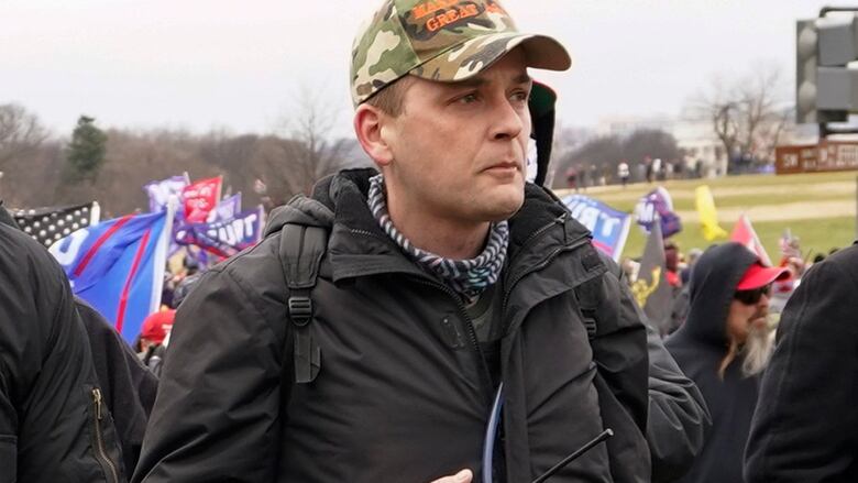 Man in camo cap and dark jacket, holding what looks like a walkie talkie, infront of a gathered group of people with flags and banners. 