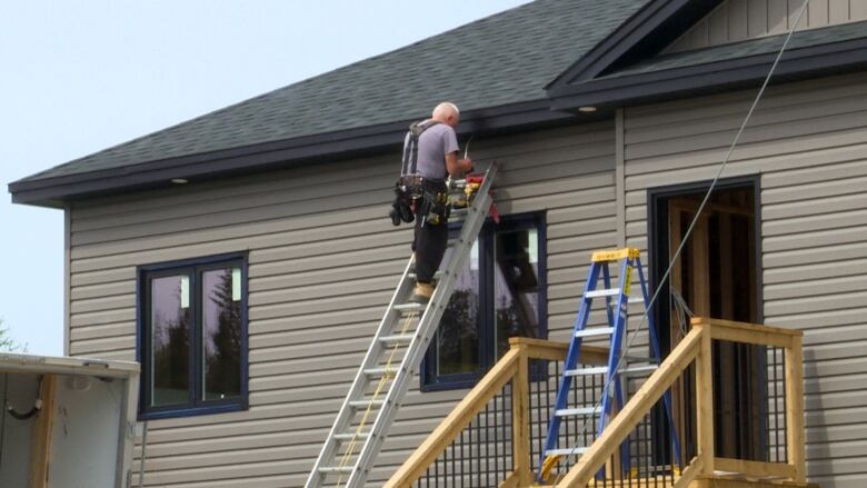 A man stands on a ladder that is leaned against the side of a house. He is putting in a pot light on the outside. The house is visibly still under construction.