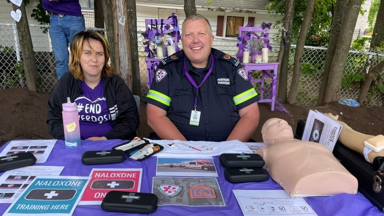 man and woman sitting at table outside with sign offering Naloxone kits