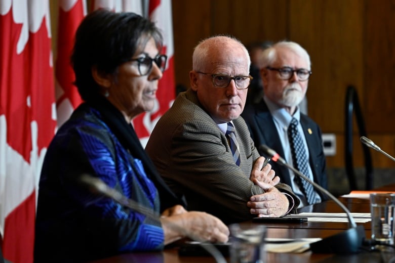 Two men sit at a desk and listen to a woman to their right speaks into a microphone.