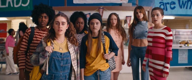 Roughly half a dozen high school girls stand together, looking off camera into the near distance. 