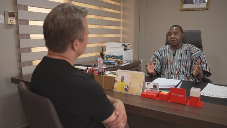 A man with light hair sits across from a man wearing a striped shirt as he sits behind a desk and speaks, gesturing with his hands. 