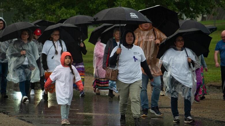 People of all ages with umbrellas walk on a residential street.