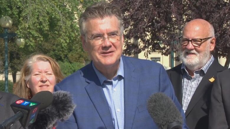 A man in glasses and short grey hair wears a blue suit and speaks from a podium outside.