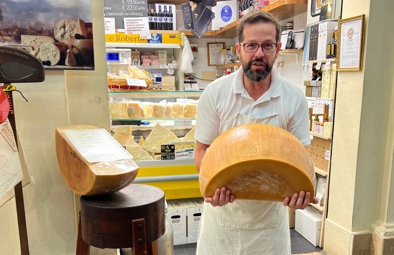 A man wearing white, with a beard and glasses, holds a large wheel of cheese.