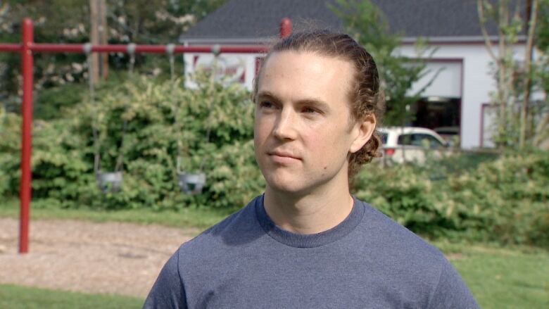 A white man with long blonde hair pulled back in a bun stands outside. A playground is seen behind him