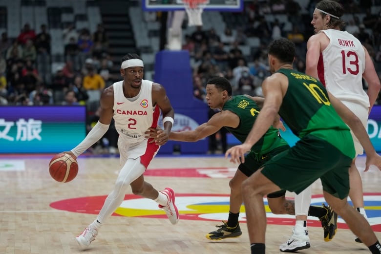 A male basketball player wearing white Canadian gear and a headband dribbles with the ball in his right hand as an opposition player touches him with one hand in an effort to guard him.