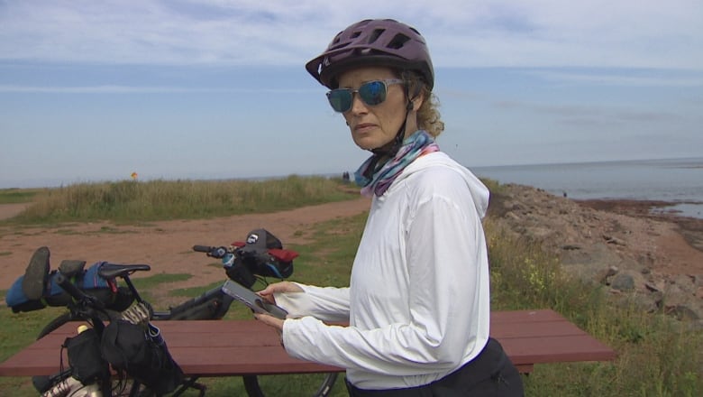 Woman wearing a bike helmet and sunglasses checks her phone in front of a scenic cliff, with her bicycle standing nearby.