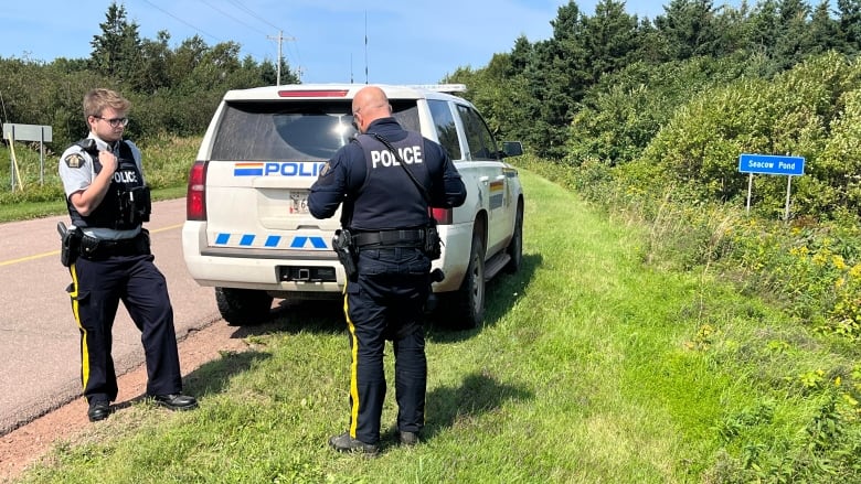 Two RCMP officers stand near an RCMP vehicle on the side of a road, with a road sign reading 'Seacow Pond' nearby.