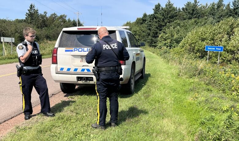 Two RCMP officers stand near an RCMP vehicle on the side of a road, with a road sign reading 'Seacow Pond' nearby.